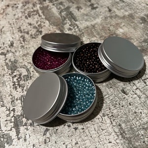 May include: Four small silver containers with lids, each filled with colorful beads. The beads are in shades of purple, brown, blue, and teal.