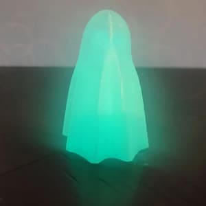 May include: A glowing green ghost-shaped night light. The light is made of a translucent plastic material and has a rounded top and a flowing bottom.
