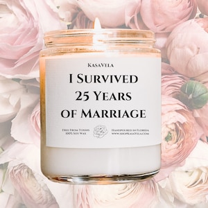 Peut inclure: Une bougie blanche avec une étiquette noire qui indique "KASAVELA I SURVIVED 25 YEARS OF MARRIAGE FREE FROM TOXINS 100% SOY WAX HANDPOURED IN FLORIDA www.SHOPKASAVELA.COM". La bougie est entourée de fleurs roses.