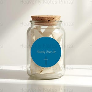 May include: A clear glass jar with a cork lid, filled with folded white paper. A blue circular label on the jar reads "Heavenly Prayer Jar" with a cross symbol. The jar is on a white surface.