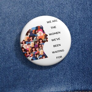 以下が含まれることがあります： 「WE ARE THE WOMEN WE'VE BEEN WAITING FOR」のテキストが入った白いボタンピン。カラフルな顔で埋められた女性の頭のシルエットが特徴です。青いデニムジャケットの上にピンが配置されています。