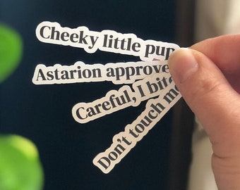 Peut inclure: Une main tenant quatre autocollants blancs avec le texte "Cheeky little pup", "Astarion approve", "Careful, I bite", et "Don't touch me".