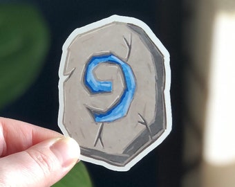 Peut inclure: Un autocollant représentant une pierre grise avec un motif de tourbillon bleu. L'autocollant a une bordure blanche et est présenté sur un fond de plante et de fenêtre avec un motif géométrique.