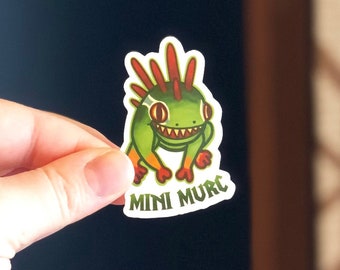 Peut inclure: Un autocollant de dessin animé vert et rouge représentant une petite créature hérissée souriante avec le texte "MINI MURE" en dessous.