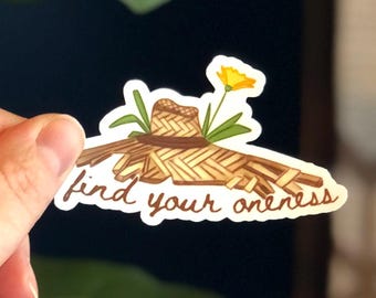 Peut inclure: Autocollant représentant un chapeau de paille avec une fleur jaune et l'inscription "find your oneness" dans une police manuscrite. L'autocollant a une bordure blanche et est posé sur un fond sombre.