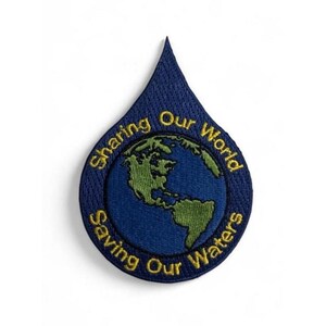 Puede incluir: Un parche bordado en forma de gota con un fondo azul oscuro y un globo terráqueo verde en el centro. El texto "Sharing Our World" está bordado en amarillo en la parte superior y "Saving Our Waters" en amarillo en la parte inferior.