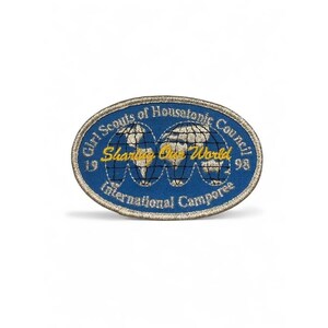 Puede incluir: Un parche bordado de forma ovalada con fondo azul y borde dorado. Presenta un gráfico de globo terráqueo con líneas de cuadrícula negras, el texto "Girl Scouts of Housatonic Council", "Sharing Our World", "International Camporee" y "1998".