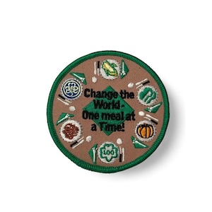 Puede incluir: Un parche bordado circular con fondo beige y borde verde. El texto "Change the World - One meal at a Time!" está en negro. Presenta ilustraciones de platos con comida, incluyendo maíz, una calabaza y varios símbolos decorativos.