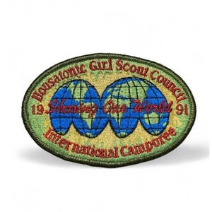 Może przedstawiać: Owalna haftowana naszywka na jasnym zielonym tle przedstawiająca dwa niebieskie globusy z zielonymi kontynentami. Napis głosi Housatonic Girl Scout Council, 1991, Sharing Our World oraz International Camporee czerwoną i ciemnozieloną nicią.