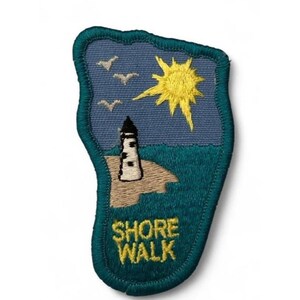 May include: An embroidered patch with a teal border featuring a blue sky, a bright yellow sun, and three white birds. A black and white lighthouse stands on a tan sandy shore next to dark teal water. The text SHORE WALK is stitched in yellow.