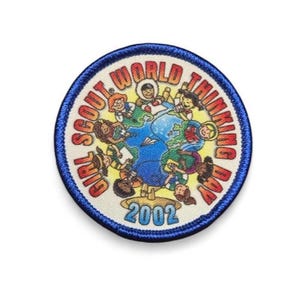 May include: A circular embroidered patch with a blue border featuring a graphic of children from diverse backgrounds surrounding a globe. The text reads Girl Scout World Thinking Day 2002 in red and blue lettering against a cream background.
