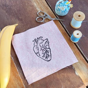 May include: A pink linen square with a black embroidered anatomical heart design. Beside it are sewing tools: silver scissors, a blue floral pin cushion, and spools of blue thread. A yellow banana is in the corner.