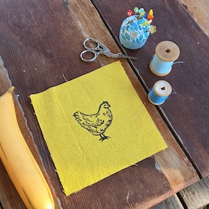 May include: A yellow fabric square with a black chicken illustration, scissors, a blue floral pin cushion with pins, and two spools of blue thread. A banana is in the lower left corner. The items are arranged on a weathered wooden surface.