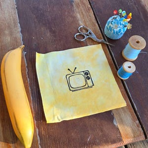 May include: A still-life arrangement on a wooden surface features a yellow banana, a yellow fabric square with a black TV graphic, blue thread spools, a blue floral pin cushion with colorful pins, and small silver scissors. The items are arranged on a rustic wooden table.