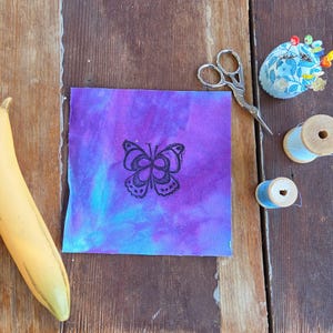 May include: A square of purple and blue fabric with a black butterfly design, next to sewing tools including scissors, a pin cushion, and spools of thread. A yellow banana is in the corner.