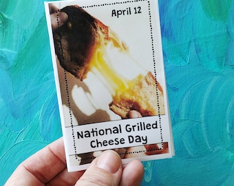 May include: A small card held in a hand, featuring a photo of a grilled cheese sandwich with melted cheese. The card has the text "April 12" and "National Grilled Cheese Day" in black text on a white background. The background is a mix of blue and green.