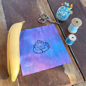 May include: A still-life image featuring a yellow banana, a piece of fabric with a sleeping bear design, sewing tools, and spools of thread. The fabric has a tie-dye effect in shades of purple and blue. A blue floral pin cushion and small scissors are also present.