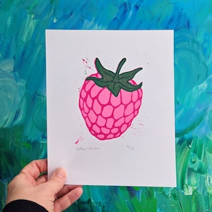 May include: A pink raspberry print on white paper with green accents. The raspberry is the focal point, with a textured design and green leaves. The background is a vibrant blue and green abstract painting. The print is signed and numbered.