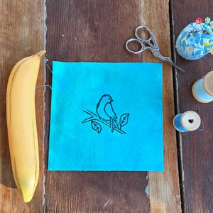 May include: A still-life arrangement on a wooden surface features a yellow banana, a teal fabric square with a black bird drawing, and sewing tools. The tools include scissors, a pin cushion, and spools of blue thread.