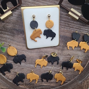 May include: A collection of handmade earrings featuring buffalo silhouettes in black and gold. The earrings are made of polymer clay and have a minimalist design.