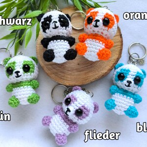 Könnte beinhalten: Gehäkelte Panda-Bären-Schlüsselanhänger in verschiedenen Farben: Schwarz, Orange, Grün, Lila und Blau. Jeder Panda hat große schwarze Augen und einen silbernen Schlüsselring. Die Schlüsselanhänger werden auf einer Holzscheibe präsentiert.