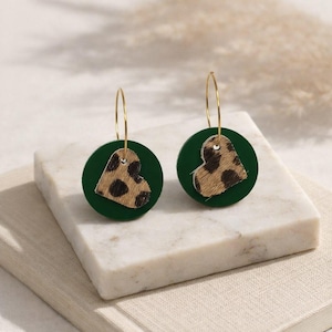 May include: A pair of gold hoop earrings. Each earring has a dark green disc with a heart-shaped patch of animal print fur. The earrings are displayed on a white marble surface.