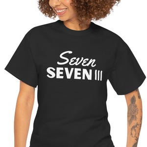 May include: Black t-shirt with the white text "Seven SEVEN III". The shirt is a classic crew neck style. The person in the image is wearing blue jeans.