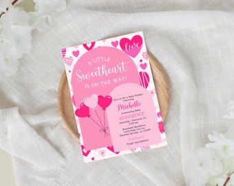 Puede incluir: Una invitación rosa para baby shower con el texto "A Little Sweetheart is on the Way!" y globos en forma de corazón. La invitación incluye detalles del evento y está sobre una bandeja de madera.