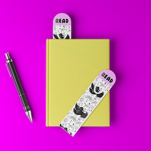 May include: A yellow book with two decorative bookmarks and a black pen on a bright pink background. The bookmarks have a pink top with the word "READ" and a black and white illustration. One bookmark is inside the book.