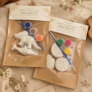 May include: Two craft kits labelled PAINT YOUR OWN MAGNETS in clear plastic bags with brown paper headers. Each kit contains white plaster shapes, a small paintbrush, and three small paint pots. One kit features a dinosaur and dolphin, the other fruit shapes.