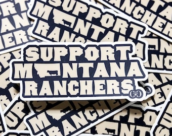 Puede incluir: Una pila de pegatinas de color marrón claro y negro con el texto "Support Montana Ranchers" en letras negras y negritas. Las pegatinas presentan una silueta del estado de Montana con una vaca en su interior.