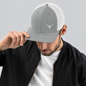 May include: A gray and white trucker hat with a white embroidered bull's head logo. The hat has a mesh back and a curved brim.