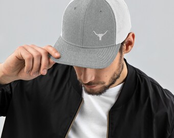 Może przedstawiać: Szara i biała czapka trucker z haftowanym białym logo głowy byka. Czapka ma siateczkowe plecy i zakrzywiony daszek.