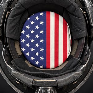 May include: A black tactical helmet interior featuring a circular crown pad printed with the American flag pattern, including white stars on a blue field and red and white stripes, surrounded by black padded lining and a rigid outer shell.