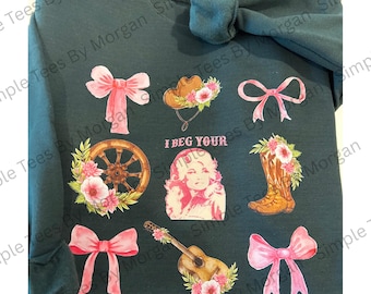 May include: Dark teal sweatshirt featuring various country-themed graphics. Includes pink bows, a cowboy hat, a wagon wheel, a guitar, a cowboy boot, and a portrait of a woman. The text "I BEG YOUR" is also visible.