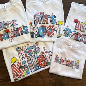 May include: Five white t-shirts with colorful, cartoon-style lettering. The shirts feature the names "Mrs. Hudgins", "Mrs. Root", "Mrs. Barnes", "Mrs. Miller", and "Huntleigh".