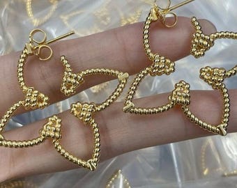 Puede incluir: Un par de pendientes de aro dorados con un diseño anudado. Los pendientes están hechos de un material trenzado similar a una cuerda.