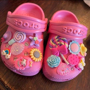 May include: Pink Crocs decorated with colorful charms, including a rainbow, a mermaid, a sunflower, a lollipop, and a seashell.