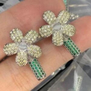 May include: A pair of silver flower-shaped earrings with green stems. Each flower is adorned with clear and yellow cubic zirconia stones.