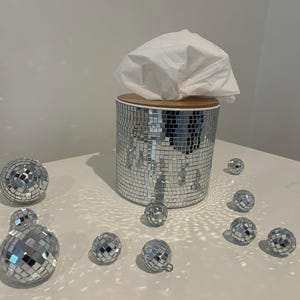 May include: A silver disco ball tissue box with a wooden lid, surrounded by several small disco balls on a white surface. The tissue box is covered in mirrored tiles, reflecting light. A white tissue is visible.