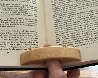 May include: A wooden finger reading aid, also known as a page holder, is shown resting on an open book. The page holder is light brown and has a curved shape. The book is open to a page with text about the history of trade.