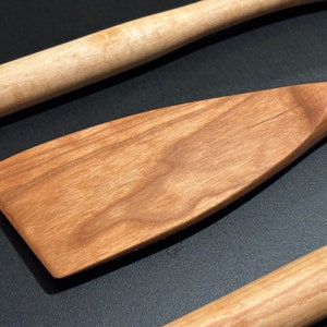 May include: Three wooden kitchen utensils: a rolling pin, a wooden spatula, and a wooden spoon. The utensils are made of light brown wood and have a natural finish.