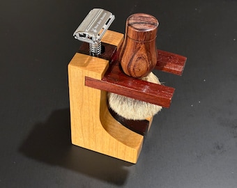 Può includere: Un supporto in legno con un pennello da barba, un rasoio e un contenitore in legno. Il supporto è in legno chiaro con accenti in legno scuro. Il rasoio è un rasoio di sicurezza argentato con la scritta "Van Der Hagen" su di esso.