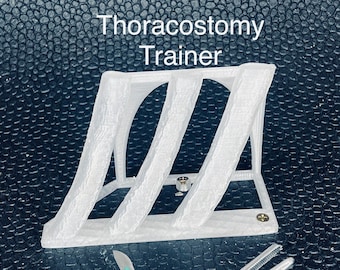 以下が含まれることがあります： 白いプラスチック製の胸腔ドレナージトレーナーモデルで、その上に「Thoracostomy Trainer」というテキストがあります。モデルは、黒いテクスチャのある表面に置かれ、その横に2つの外科用器具があります。