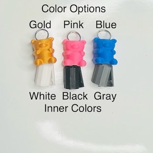 Op de afbeelding: Drie gummy bear-bedels met verschillende kleuren lichamen en binnenkleuren. De gummy bears zijn goud, roze en blauw. De binnenkleuren zijn wit, zwart en grijs. De bedels zijn bevestigd aan zilveren ringen.