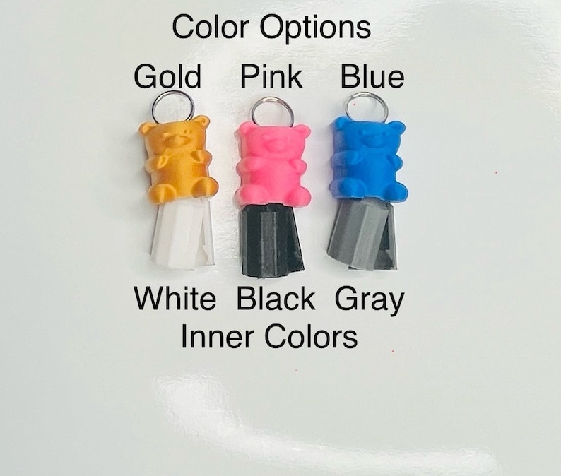 Op de afbeelding: Drie gummy bear-bedels met verschillende kleuren lichamen en binnenkleuren. De gummy bears zijn goud, roze en blauw. De binnenkleuren zijn wit, zwart en grijs. De bedels zijn bevestigd aan zilveren ringen.