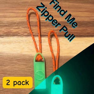 Puede incluir: Dos tiradores de cremallera que brillan en la oscuridad con cordones naranjas. Los tiradores de cremallera son verdes y tienen un pequeño logotipo. El texto "Find Me Zipper Pull" está escrito en blanco sobre fondo negro. El texto "2 pack" está escrito en amarillo sobre fondo negro.