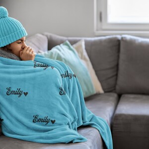 May include: A person wrapped in a blue fleece blanket with the name "Emily" printed on it, sitting on a grey sofa. The person is wearing a blue knitted hat with a white pom-pom.