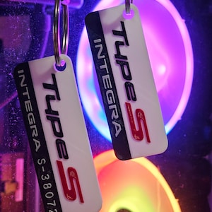 May include: Two white rectangular keychains with the text "INTEGRA Type S" in black and red. The keychains are attached to silver keyrings and hang against a background of colorful lights.