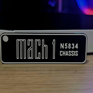 May include: A black rectangular keychain with the text "MACH 1" in a white, stylized font. Below the text is "N5834 CHASSIS" in white. The keychain has a small hole on the left side.